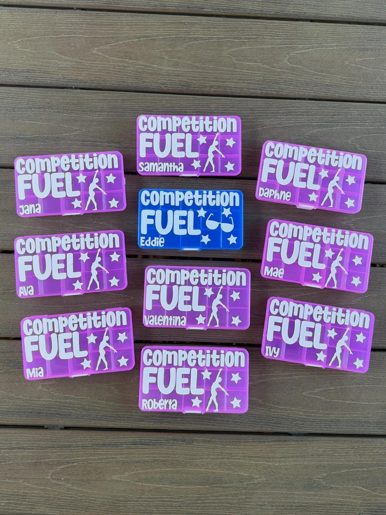 Pode incluir: Uma cole&ccedil;&atilde;o de nove recipientes retangulares de pl&aacute;stico para lanches em roxo e azul. Cada recipiente apresenta o texto branco "competition FUEL" com uma silhueta de ginasta e estrelas. Nomes individuais est&atilde;o impressos em cada um, incluindo Jana, Samantha, Daphne, Ava, Eddie, Mae, Valentina, Mia e Roberta.
