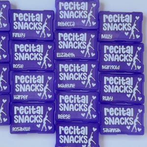 Pode incluir: Uma cole&ccedil;&atilde;o de recipientes retangulares de pl&aacute;stico roxo para lanches, cada um com o texto branco "recital SNACKS", a silhueta de um dan&ccedil;arino e dois cora&ccedil;&otilde;es. Cada recipiente &eacute; personalizado com um nome diferente, incluindo Rebecca, Finley, Milly, Rosie, Marigold, Harper, Madeline, Ruby, Rosabelle, Reese, Savannah e Abigail.