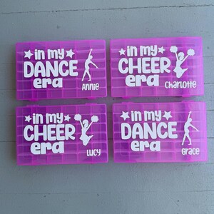 May include: Four magenta plastic storage boxes with white text and graphics. Two boxes say "in my DANCE era" with a dancer silhouette and the names "Annie" and "Grace". The other two boxes say "in my CHEER era" with a cheerleader graphic and the names "Charlotte" and "Lucy".