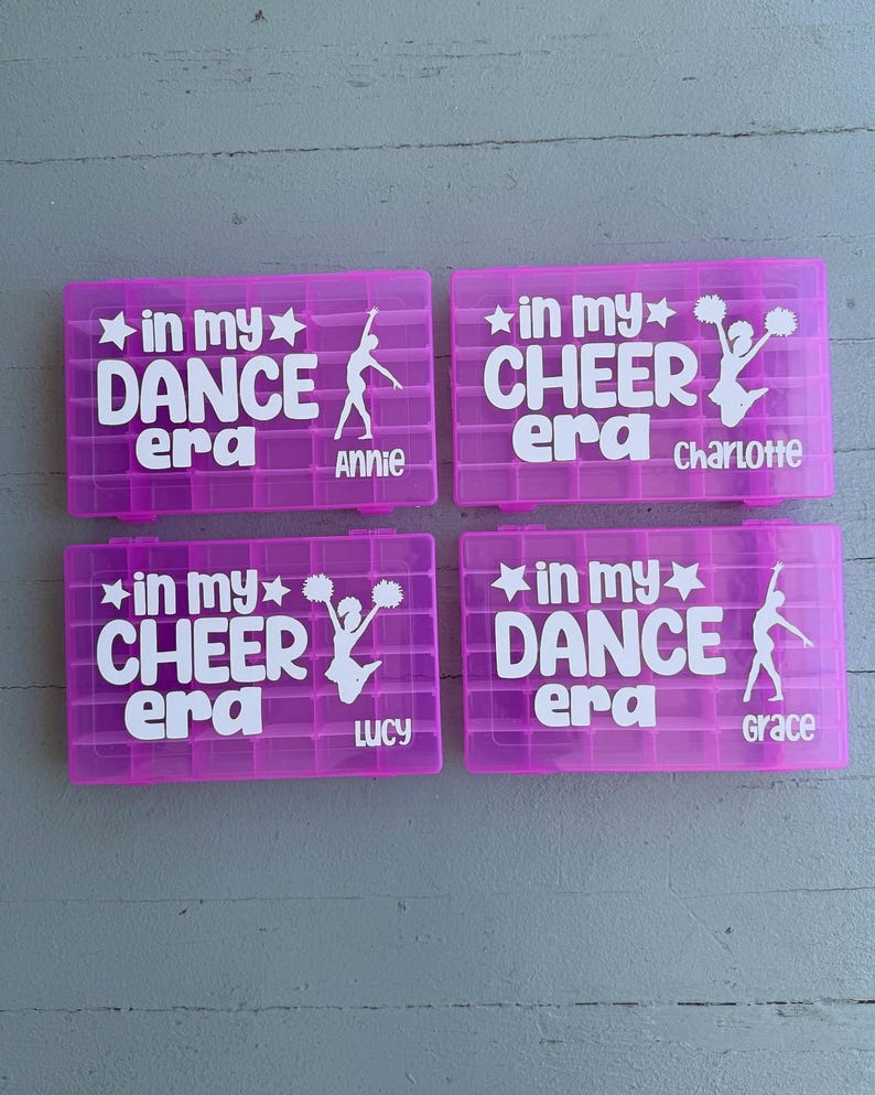 May include: Four magenta plastic storage boxes with white text and graphics. Two boxes say "in my DANCE era" with a dancer silhouette and the names "Annie" and "Grace". The other two boxes say "in my CHEER era" with a cheerleader graphic and the names "Charlotte" and "Lucy".