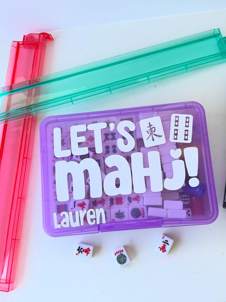 May include: A purple Mahjong set with the text "LET'S mahj!" and the name "Lauren" in white. The set includes tiles, dice, and a purple case. Two clear plastic trays are visible in pink and green.