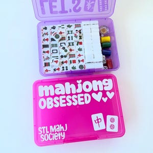 May include: A purple and pink Mahjong set in two plastic cases. The top case is purple and open, revealing tiles, dice, and other game pieces. The bottom case is pink and has the words "mahjong OBSESSED" and "STL Mahj Society" printed on it.