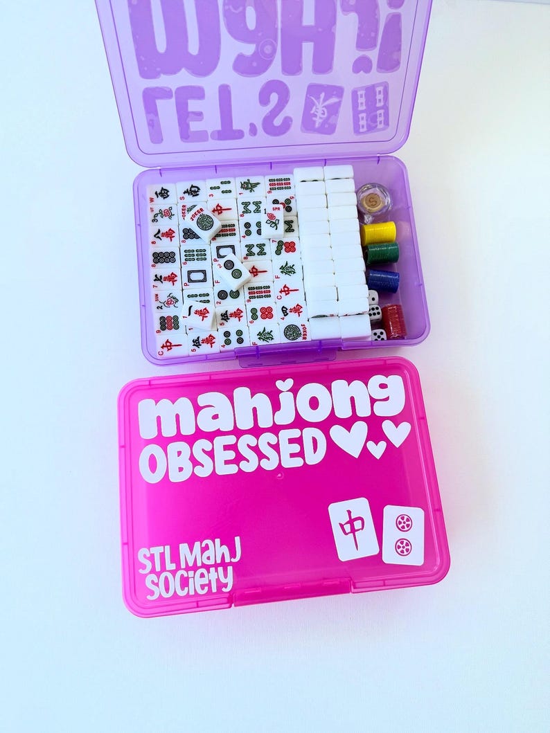 May include: A purple and pink Mahjong set in two plastic cases. The top case is purple and open, revealing tiles, dice, and other game pieces. The bottom case is pink and has the words "mahjong OBSESSED" and "STL Mahj Society" printed on it.