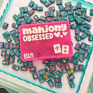 May include: A pink Mahjong tile case with the words "mahjong OBSESSED" in white, surrounded by teal Mahjong tiles. The case is personalized with the name "ELLIS". The tiles feature various symbols and numbers.