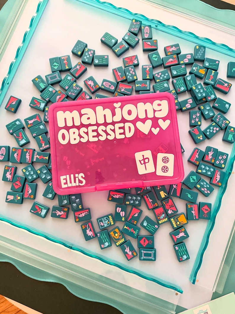 May include: A pink Mahjong tile case with the words "mahjong OBSESSED" in white, surrounded by teal Mahjong tiles. The case is personalized with the name "ELLIS". The tiles feature various symbols and numbers.