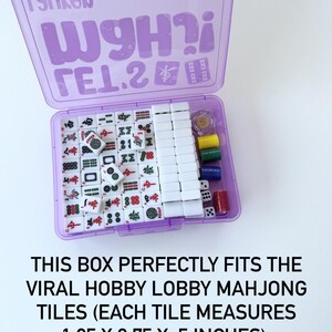 May include: A purple plastic box containing a Mahjong set. The box is open, revealing white tiles with red, green, and black symbols. Dice and colored chips are also visible. The text on the box reads "TSOLGN WgH?i &Gamma;&Epsilon;1.2".