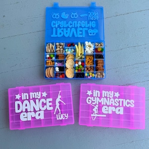 May include: A blue snack box filled with various snacks, and two pink boxes with white text that reads "in my DANCE era Lucy" and "in my GYMNASTICS era". The blue box has text on the lid.