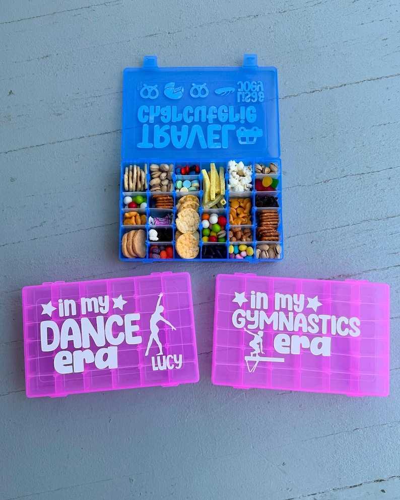 May include: A blue snack box filled with various snacks, and two pink boxes with white text that reads "in my DANCE era Lucy" and "in my GYMNASTICS era". The blue box has text on the lid.