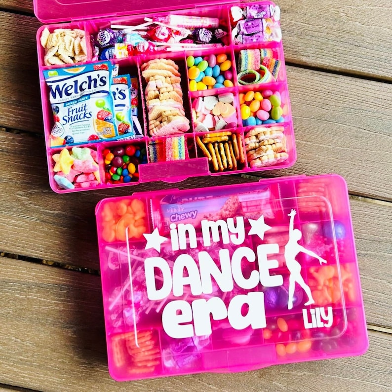 May include: Two pink plastic divided boxes filled with sweets, biscuits, and snacks. The bottom box has the text "in my DANCE era" with a dancer silhouette and the name "Lily". The top box is filled with a variety of colourful treats.