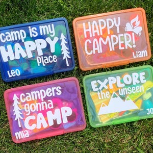 May include: Four clear plastic containers with colourful lids and personalised text. The containers have different coloured lids: blue, orange, pink, and green. The text on the containers reads: "Camp is my Happy Place", "Happy Camper", "Campers gonna Camp", and "Explore the unseen".