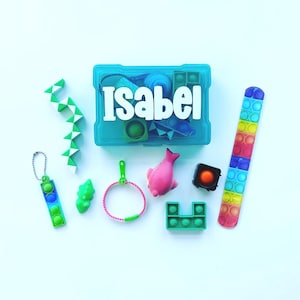 Könnte beinhalten: Eine türkisfarbene Kunststoff-Aufbewahrungsbox mit dem Namen Isabel in weißen Buchstaben, umgeben von verschiedenen sensorischen Spielzeugen, darunter ein grün-weißes Schlangenpuzzle, ein regenbogenfarbenes Pop-it-Armband, ein rosa Delfin und weitere kleine Fidget-Artikel.