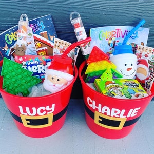 May include: Two red Santa-themed gift buckets filled with Christmas-themed toys and treats. Each bucket has a name on it, one says "LUCY" and the other says "CHARLIE". The buckets contain coloring books, pop-it toys, and Santa and snowman-shaped cups.
