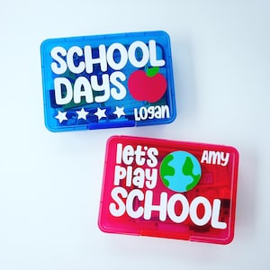 May include: Two clear plastic pencil boxes with colorful designs and text. The blue box has a red apple and the text "SCHOOL DAYS" with the name "Logan". The pink box has a blue globe and the text "Let's Play SCHOOL" with the name "Amy".