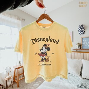 Op de afbeelding: Een lichtgeel t-shirt met korte mouwen en de tekst Disneyland California en Est 1955 in donkerbruine letters. Een afbeelding van Minnie Mouse met een rode rok met stippen en een bloemenhoed staat centraal op de voorzijde van het kledingstuk.