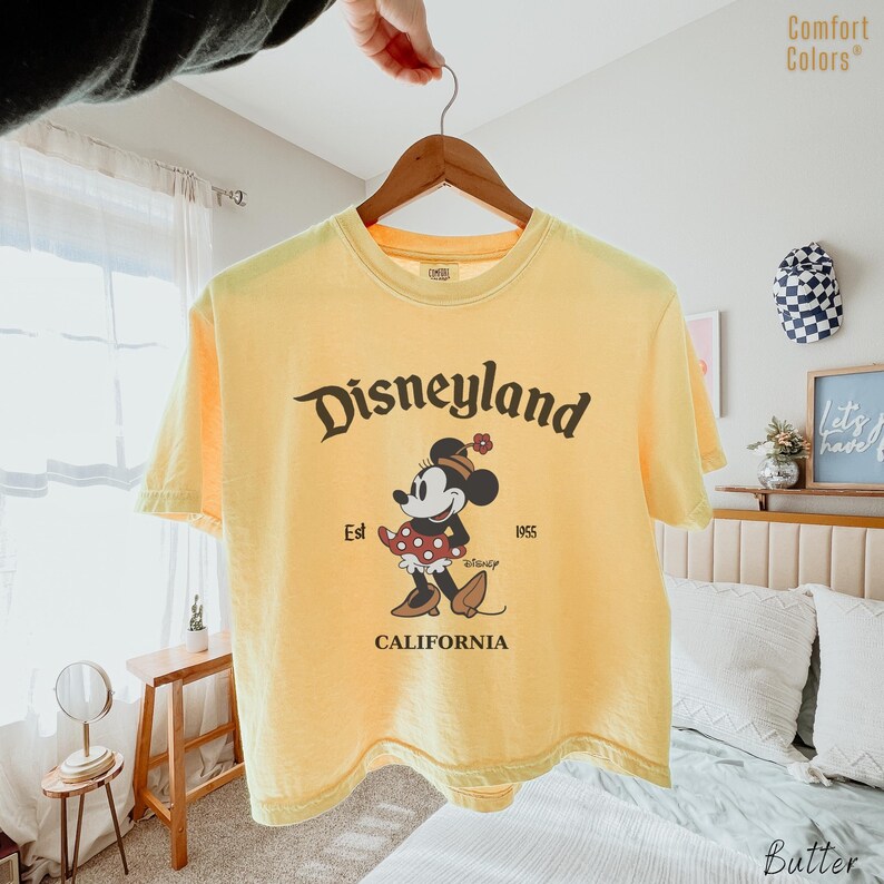 Op de afbeelding: Een lichtgeel t-shirt met korte mouwen en de tekst Disneyland California en Est 1955 in donkerbruine letters. Een afbeelding van Minnie Mouse met een rode rok met stippen en een bloemenhoed staat centraal op de voorzijde van het kledingstuk.