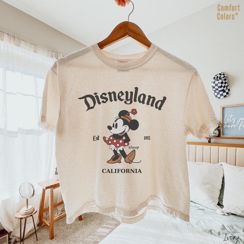 Op de afbeelding: Een ivoorkleurig T-shirt met korte mouwen en een afbeelding van Minnie Mouse met een hoed en een rood jurkje met stippen. De tekst Disneyland, Est 1955 en California staat in een donkerbruin lettertype. Het shirt hangt aan een houten hanger.