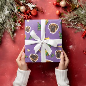 May include: A wrapped gift with a purple background featuring Mastiff dog faces, Christmas trees, and stars. The gift is tied with a white ribbon and bow. The background is a festive red with Christmas greenery.