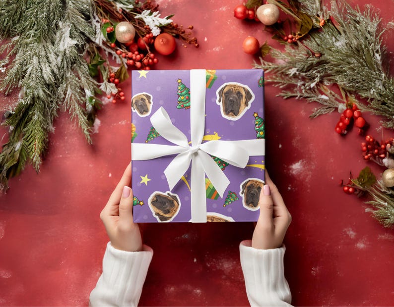 May include: A wrapped gift with a purple background featuring Mastiff dog faces, Christmas trees, and stars. The gift is tied with a white ribbon and bow. The background is a festive red with Christmas greenery.