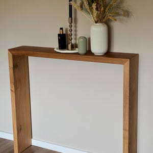 Puede incluir: Una consola de madera con acabado natural. La mesa es rectangular y de diseño minimalista. Está decorada con una vela negra, un candelabro dorado, un jarrón blanco con plantas secas, un pequeño jarrón verde y un frasco de perfume.