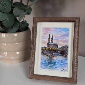 Puede incluir: Una pintura de acuarela enmarcada de una catedral y un puente sobre un río, contra un cielo colorido. La obra de arte está en un marco de madera marrón, exhibida sobre una superficie blanca, con una planta en maceta en el fondo.