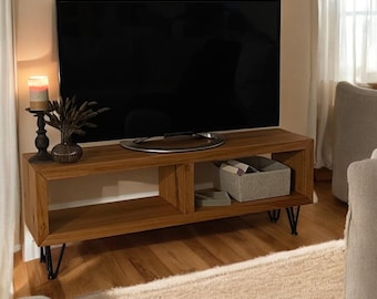 May include: A wooden TV stand with black hairpin legs. The stand has two shelves and is holding a TV, a candle, and a decorative plant. The stand is in front of a beige wall and a white rug.