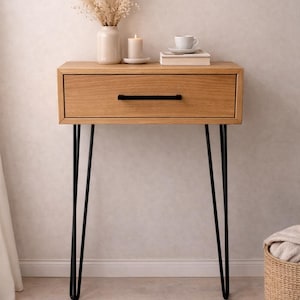 Puede incluir: Mesa auxiliar de madera clara con cajón y patas negras de horquilla. Un jarrón con flores secas, una vela, una pila de libros y una taza de té con platillo encima. Una pieza de mobiliario elegante.