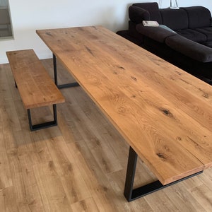 May include: A long wooden dining table with a natural wood finish and black metal legs. The table is set on a light wood floor. A matching wooden bench with black metal legs is in the foreground.