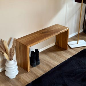 Puede incluir: Un banco de madera con un acabado natural y un diseño sencillo. El banco tiene una parte superior ancha y plana y una base estrecha y abierta. El banco está hecho de madera maciza y tiene un aspecto rústico. El banco está sentado en un suelo de madera y hay dos pares de botas negras debajo.