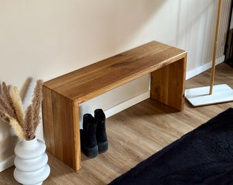 May include: A wooden bench with a natural finish and a simple design. The bench has a wide, flat top and a narrow, open base. The bench is made of solid wood and has a rustic look. The bench is sitting on a wooden floor and there are two pairs of black boots underneath.