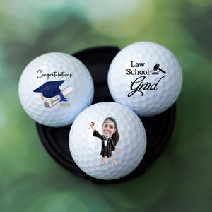 Könnte beinhalten: Drei weiße Golfbälle mit Abschlussmotiven. Einer trägt die Aufschrift "Herzlichen Glückwunsch" mit einer Abschlusskappe und einer Urkunde. Ein anderer trägt die Aufschrift "Law School Grad" mit einem Hammer. Der dritte Ball zeigt einen Cartoon-Absolventen.
