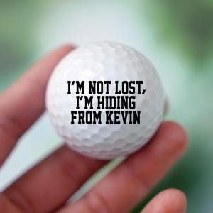Könnte beinhalten: Ein weißer Golfball mit schwarzem Text, der "Ich bin nicht verloren, ich verstecke mich vor Kevin" lautet.