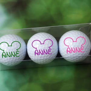 May include: Three white golf balls with a pink, green, and purple Mickey Mouse head design and the name "Anne" printed on each ball. The golf balls are in a clear plastic box.