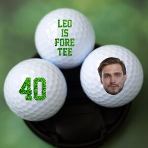 May include: Three white golf balls with green text and a face printed on one of the balls. The text on the balls reads "Leo is Fore Tee" and "40".