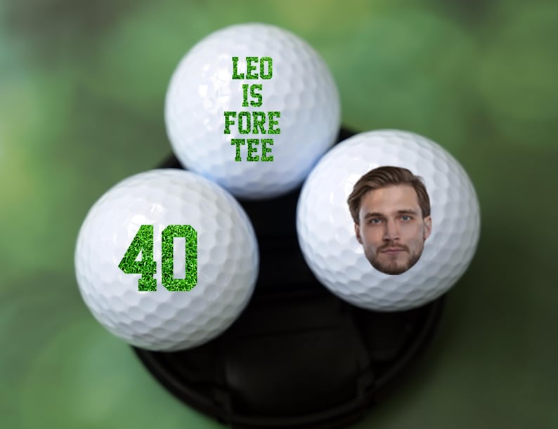 May include: Three white golf balls with green text and a face printed on one of the balls. The text on the balls reads "Leo is Fore Tee" and "40".