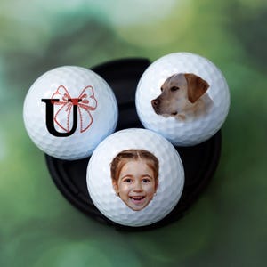 Może przedstawiać: Trzy białe piłki golfowe z personalizowanymi wzorami. Jedna piłka ma czerwoną kokardkę i literę "U" nadrukowaną na niej. Inna piłka ma zdjęcie psa rasy golden retriever. Trzecia piłka ma zdjęcie młodej dziewczyny z blond włosami.