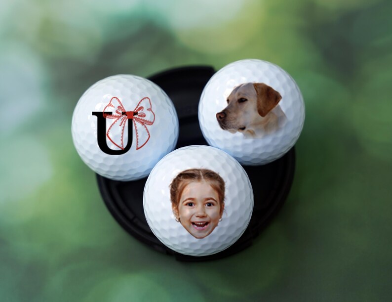 Może przedstawiać: Trzy białe piłki golfowe z personalizowanymi wzorami. Jedna piłka ma czerwoną kokardkę i literę "U" nadrukowaną na niej. Inna piłka ma zdjęcie psa rasy golden retriever. Trzecia piłka ma zdjęcie młodej dziewczyny z blond włosami.