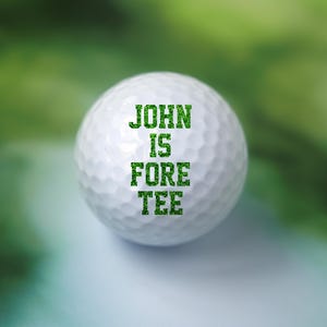 May include: A white golf ball with green text that reads "JOHN IS FORE TEE".