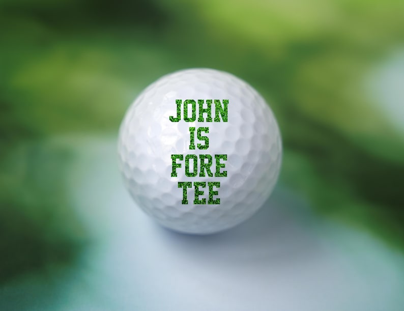 May include: A white golf ball with green text that reads "JOHN IS FORE TEE".