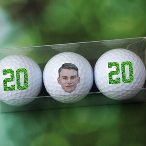 May include: Three white golf balls with green glitter numbers '20' on two of the balls. The third ball has a photo of a person's face on it.