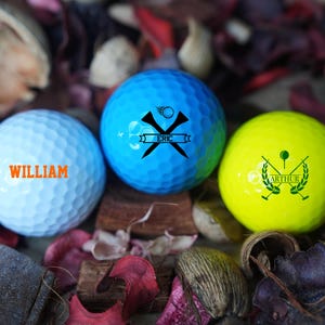 May include: Three personalized golf balls with different designs. The blue golf ball has a crossed golf club design with the name "ERIC" on a banner. The white golf ball has the name "WILLIAM" in orange text. The yellow golf ball has a crossed golf club design with a laurel wreath and the name "ARTHUR" on a banner.