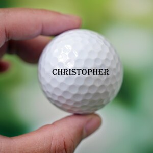 Może przedstawiać: Biała piłka golfowa z nadrukowanym imieniem "CHRISTOPHER".