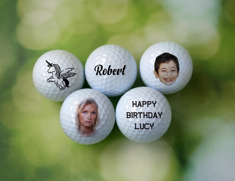 Może przedstawiać: Pięć białych piłek golfowych z personalizowanymi wzorami. Jedna piłka ma jednorożca ze skrzydłami, druga ma napis "Robert", trzecia ma twarz dziecka, czwarta ma napis "Happy Birthday Lucy", a ostatnia ma twarz kobiety.