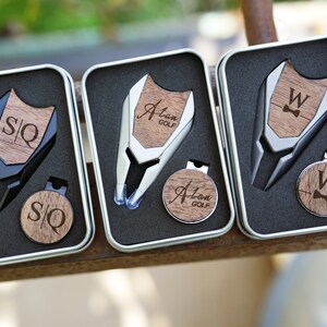 Könnte beinhalten: Drei silberne Golfball-Marker-Sets in schwarzen, silbernen und Holzkisten. Jedes Set enthält einen Golfball-Marker, ein Divot-Tool und einen magnetischen Ball-Marker-Halter. Die Ball-Marker sind mit den Initialen "S/Q", "Alan Golf" und "W" mit einer Schleife graviert.
