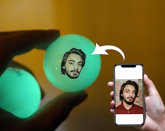 May include: Two glow-in-the-dark balls with a person's face printed on one of them. The balls are green and translucent. A smartphone is in the background showing a photo of the same person.