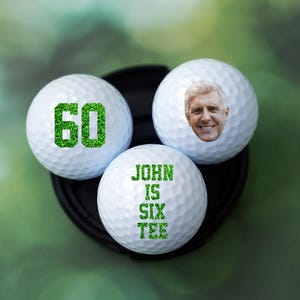 May include: Three white golf balls with green text and a photo of a man on one of the balls. The text on the balls reads "60" and "John is Six Tee".