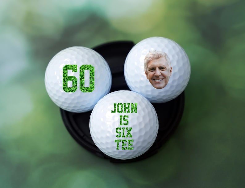 May include: Three white golf balls with green text and a photo of a man on one of the balls. The text on the balls reads "60" and "John is Six Tee".