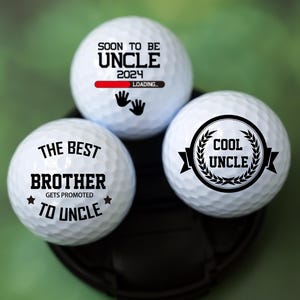 Könnte beinhalten: Drei weiße Golfbälle mit schwarzem Text. Der erste Ball sagt "Soon to be Uncle 2024 Loading." Der zweite Ball sagt "The Best Brother Gets Promoted to Uncle." Der dritte Ball sagt "Cool Uncle" in einem Kranz.