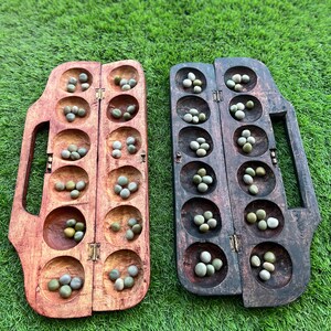 May include: A wooden game board with two rows of six holes each, filled with small green stones. The board is divided in the middle and has a handle on each side.