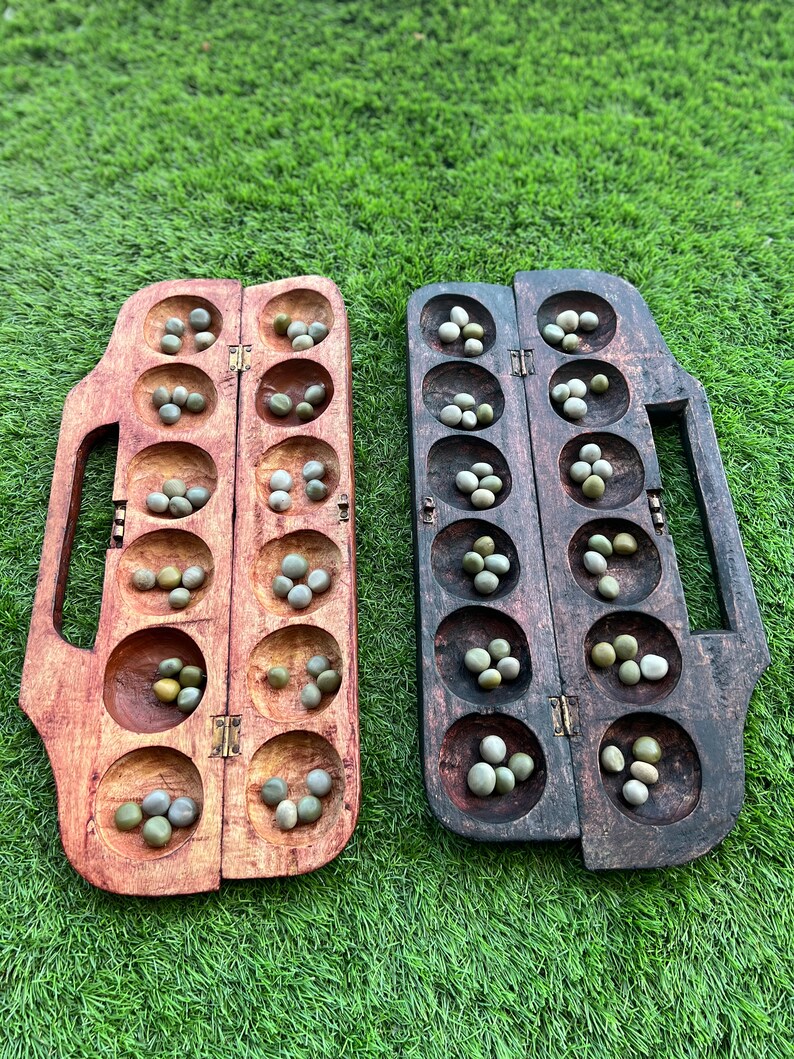 May include: A wooden game board with two rows of six holes each, filled with small green stones. The board is divided in the middle and has a handle on each side.