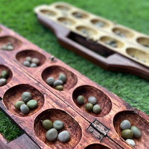 May include: A wooden game board with two rows of six depressions, each containing small green stones. The board is made of dark wood and has a metal hinge in the center.